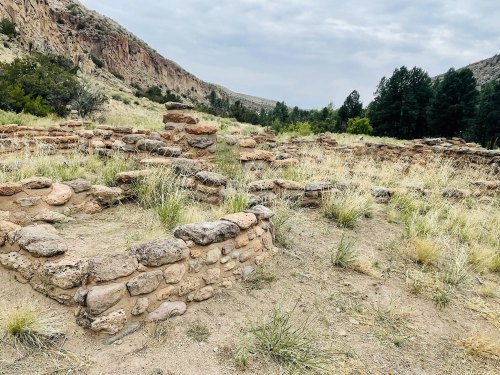 Ruins of stone structures in a grassy area, surrounded by hills and trees under a cloudy sky.