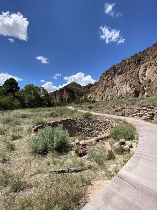 A winding path leads to a circular stone structure, surrounded by greenery and rocky cliffs under a blue sky with clouds.