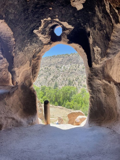 View through a cave opening, revealing a scenic landscape of trees and hills under a clear blue sky.