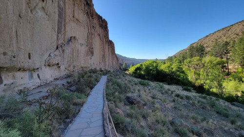 A stone pathway winds through a canyon, flanked by tall cliffs and lush greenery on one side under a clear blue sky.