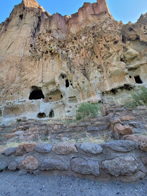 Rocky cliff face with caves and eroded features, surrounded by sparse vegetation and a stone wall in the foreground.
