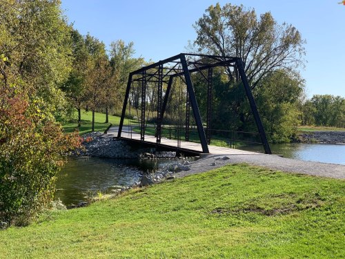 A black metal bridge spans a calm river, surrounded by green grass and trees under a clear blue sky.