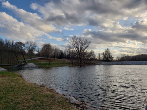 A serene lake surrounded by trees and grassy banks, with a cloudy sky reflecting on the water's surface.