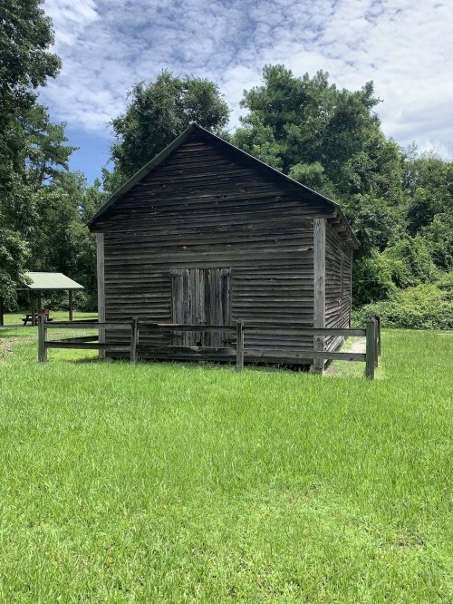 A weathered wooden cabin surrounded by lush green grass and trees under a partly cloudy sky.
