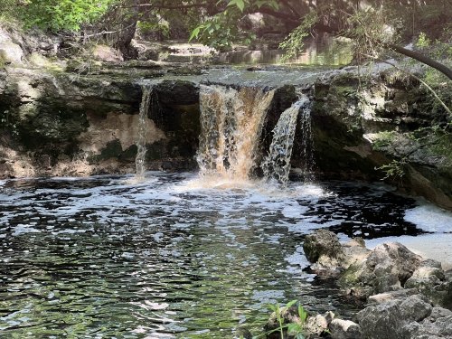 A small waterfall cascades over rocks into a dark pool, surrounded by lush greenery and natural scenery.