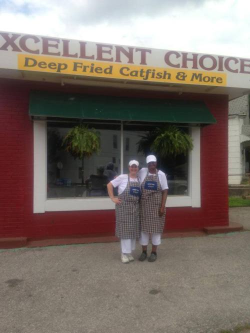 Two women in aprons stand outside a restaurant called "Excellent Choice," which specializes in deep-fried catfish.