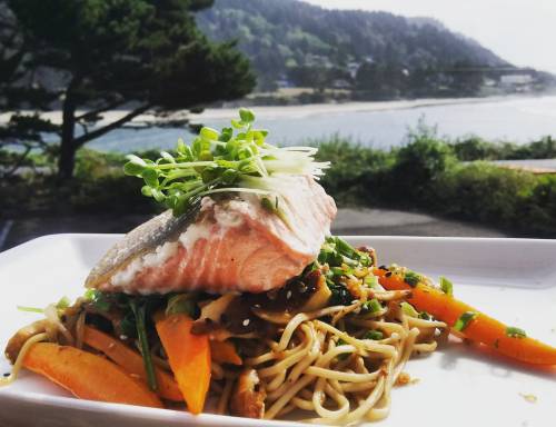 A plate of salmon atop stir-fried noodles and vegetables, with a scenic coastal view in the background.