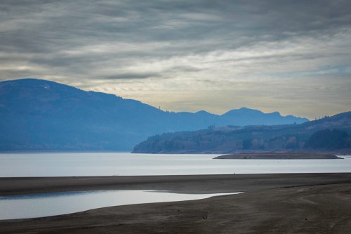 A serene landscape featuring a calm lake, distant mountains, and a cloudy sky, with a sandy shore in the foreground.