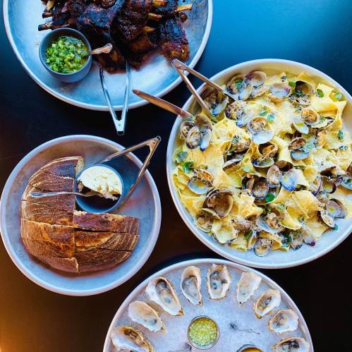 A spread of seafood pasta with clams, grilled ribs, bread, and a platter of oysters with sauce on a dark table.