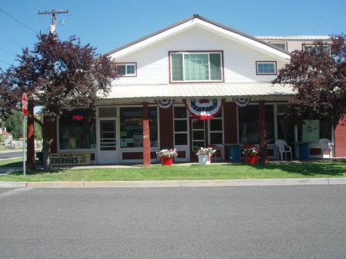 A storefront with patriotic decorations, potted flowers, and a sign for cold beverages, set in a sunny neighborhood.
