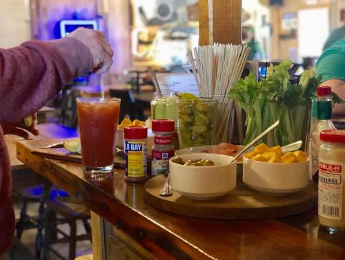 A bar counter with a cocktail, garnishes, spices, and fresh ingredients for drinks, including celery and olives.