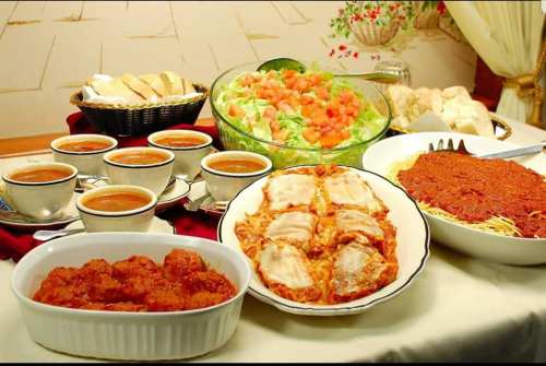 A table spread with various dishes: pasta with sauce, meatballs, salad, bread, and cups of soup.