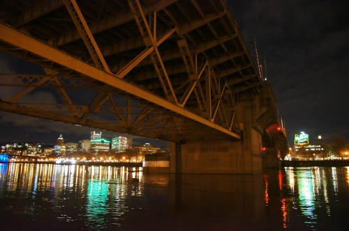 View from under a bridge at night, reflecting city lights on the water with a skyline in the background.