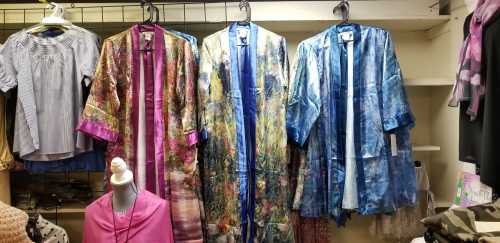 Three colorful, patterned kimonos hang on display in a clothing store, with various tops visible in the background.