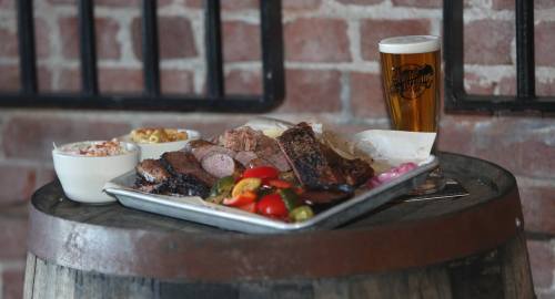 A platter of assorted meats with sides, including coleslaw and pickled vegetables, next to a glass of beer on a barrel.