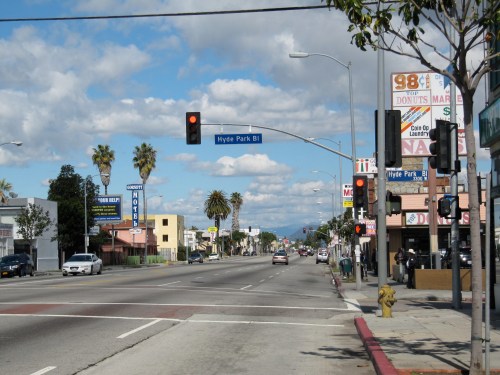 A street view featuring traffic lights, palm trees, and storefronts under a partly cloudy sky.
