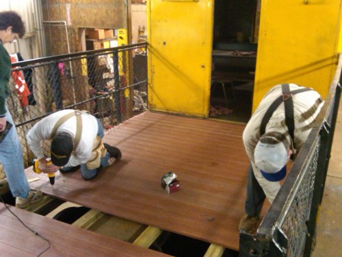Two workers installing wooden flooring on a platform, using tools in a workshop with yellow doors in the background.