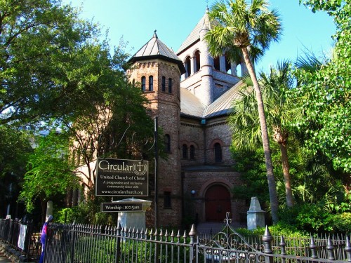 Historic brick church with a tall steeple, surrounded by trees and a decorative fence, featuring a sign for worship services.