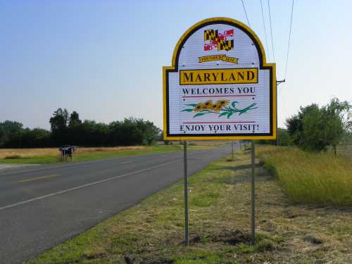 A welcome sign for Maryland along a rural road, featuring the state flag and the message "Enjoy your visit!"