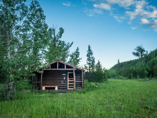 A rustic wooden cabin surrounded by lush green grass and trees under a clear blue sky.