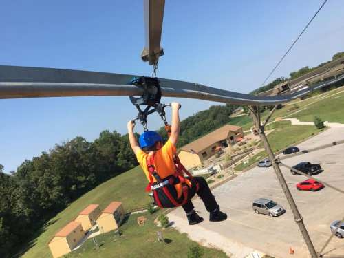 A child in a harness zip-lining above a grassy area with buildings in the background on a sunny day.