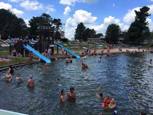 A busy swimming area with people in the water, water slides, and a sandy beach under a blue sky with clouds.