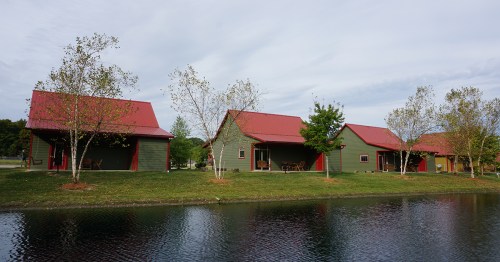 Row of green cabins with red roofs beside a calm pond, surrounded by trees and a cloudy sky.