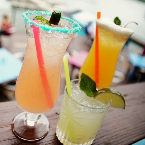 Three colorful cocktails on a wooden table, featuring vibrant garnishes and straws, with a blurred background.
