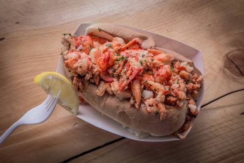 A lobster roll filled with chunks of lobster meat, garnished with herbs, served with a lemon wedge on a wooden surface.