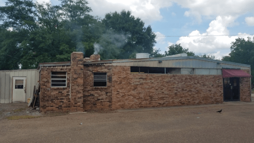 A brick building with smoke rising from the chimney, surrounded by trees and a gravel area.