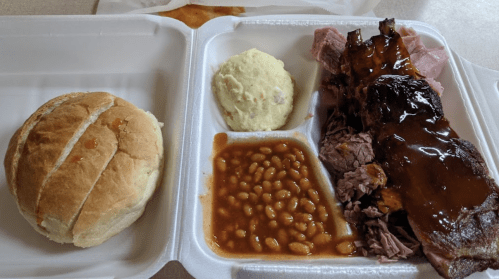 A takeout tray with ribs, pulled pork, baked beans, mashed potatoes, and a roll.