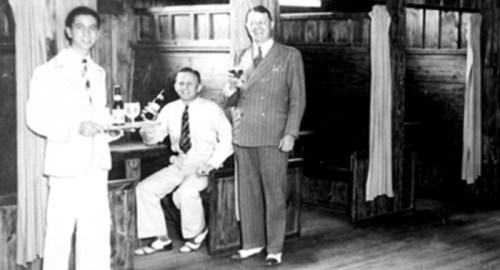 Three men in vintage attire hold drinks in a wooden interior, with one man seated and two standing.