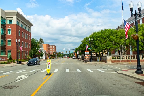 A wide street lined with trees and buildings, featuring traffic signs and American flags under a cloudy sky.
