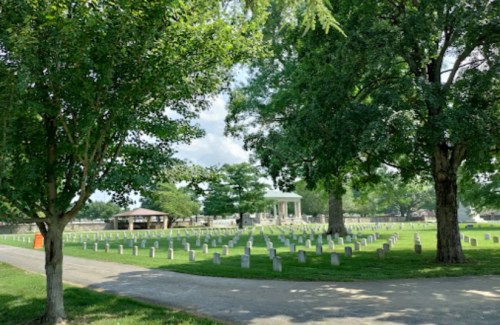 A peaceful cemetery with white gravestones, surrounded by lush green trees and a gazebo in the background.