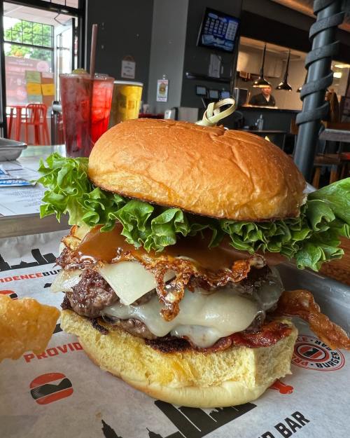 A close-up of a juicy burger with lettuce, bacon, melted cheese, and a glossy bun, served on a tray.