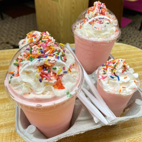 Four pink milkshakes topped with whipped cream and colorful sprinkles, served in clear cups on a tray.