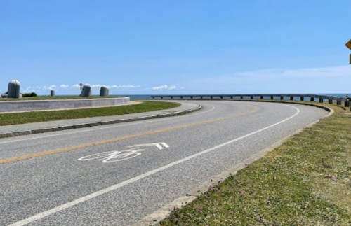 A curved road with a bike lane, leading towards the ocean under a clear blue sky. Green grass lines the roadside.