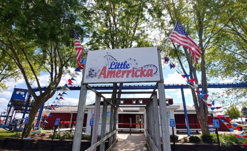 Entrance to Little Americka amusement park, decorated with flags and colorful bunting, surrounded by trees.