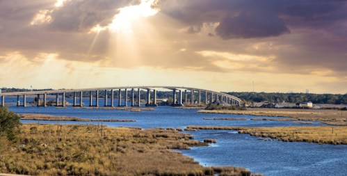 A bridge spans a river under dramatic clouds, with sunlight breaking through, surrounded by marshland and water.