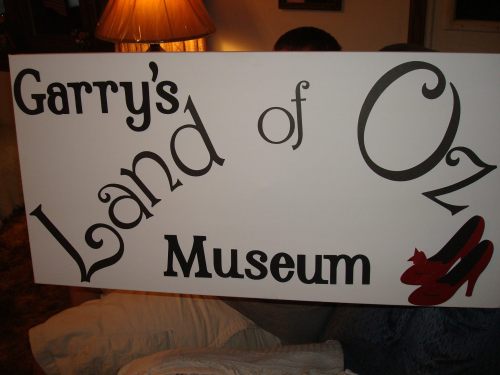 Sign for "Garry's Land of Oz Museum" featuring red shoes, with a warm background and a lamp.