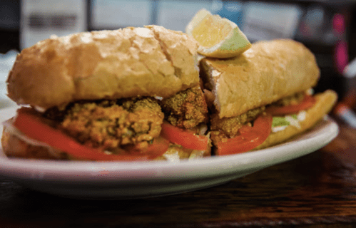 A close-up of a sandwich filled with fried seafood, lettuce, and tomato, served on a toasted bun with a lime wedge.