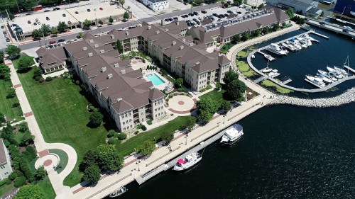 Aerial view of a waterfront hotel with a pool, marina, and landscaped grounds beside a calm body of water.