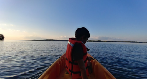 A child in a life jacket sits in a kayak on calm water, with a clear blue sky and distant trees in the background.