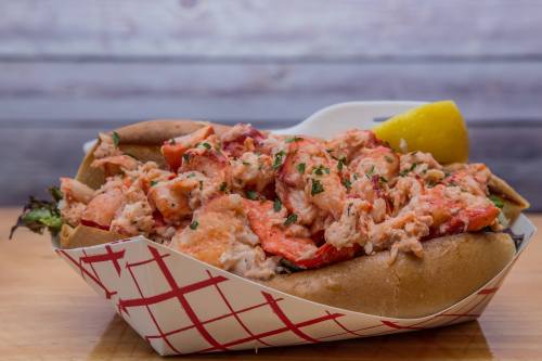 A lobster roll filled with chunks of lobster meat, garnished with parsley, served in a paper tray with a lemon wedge.