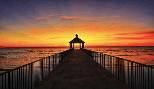 A serene sunset over the ocean, viewed from a pier leading to a small gazebo. Vibrant colors fill the sky.