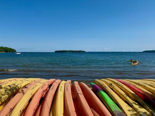 Colorful kayaks stacked on a beach, with a calm lake and distant island under a clear blue sky.