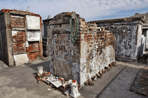 A weathered brick tomb surrounded by other crypts, adorned with colorful beads and offerings, under a cloudy sky.