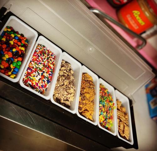 A variety of colorful toppings in small trays, including sprinkles, candies, and crushed cookies, arranged in a container.