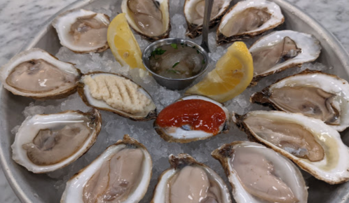 A platter of fresh oysters on ice, garnished with lemon wedges and a small cup of cocktail sauce.