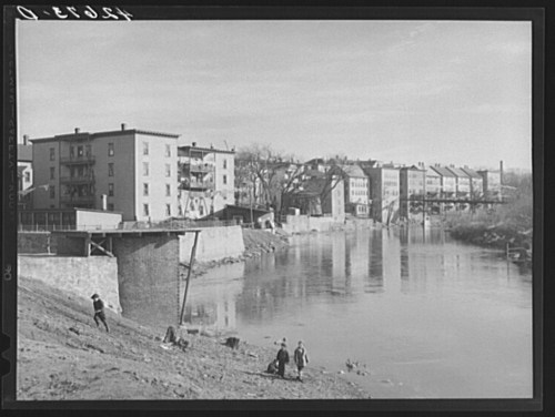 Black and white photo of a river with buildings along the bank and people walking near the water's edge.